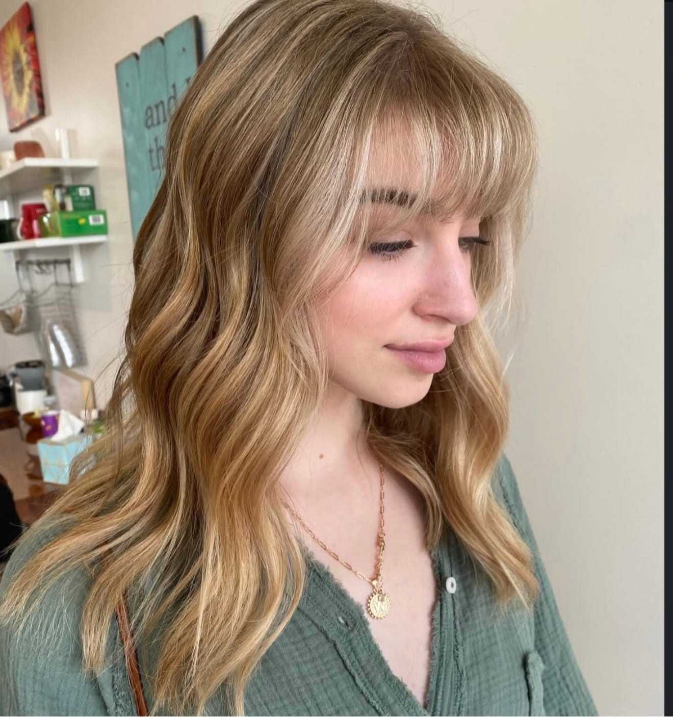 Young woman with wavy blonde hair and bangs, wearing a green top, in a cozy room setting.