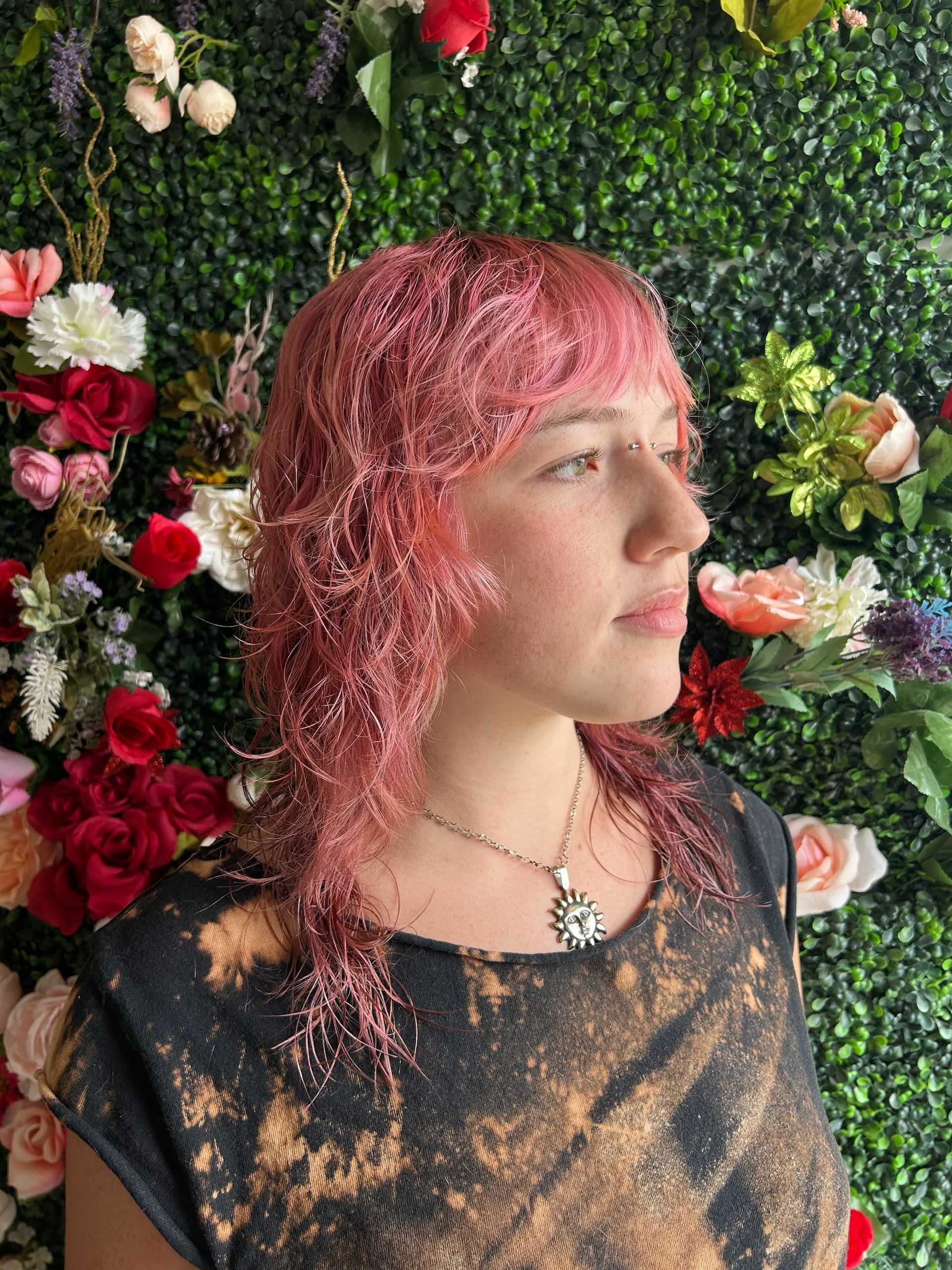 Woman with pink hair stands against floral wall, wearing tie-dye shirt and sunburst necklace.