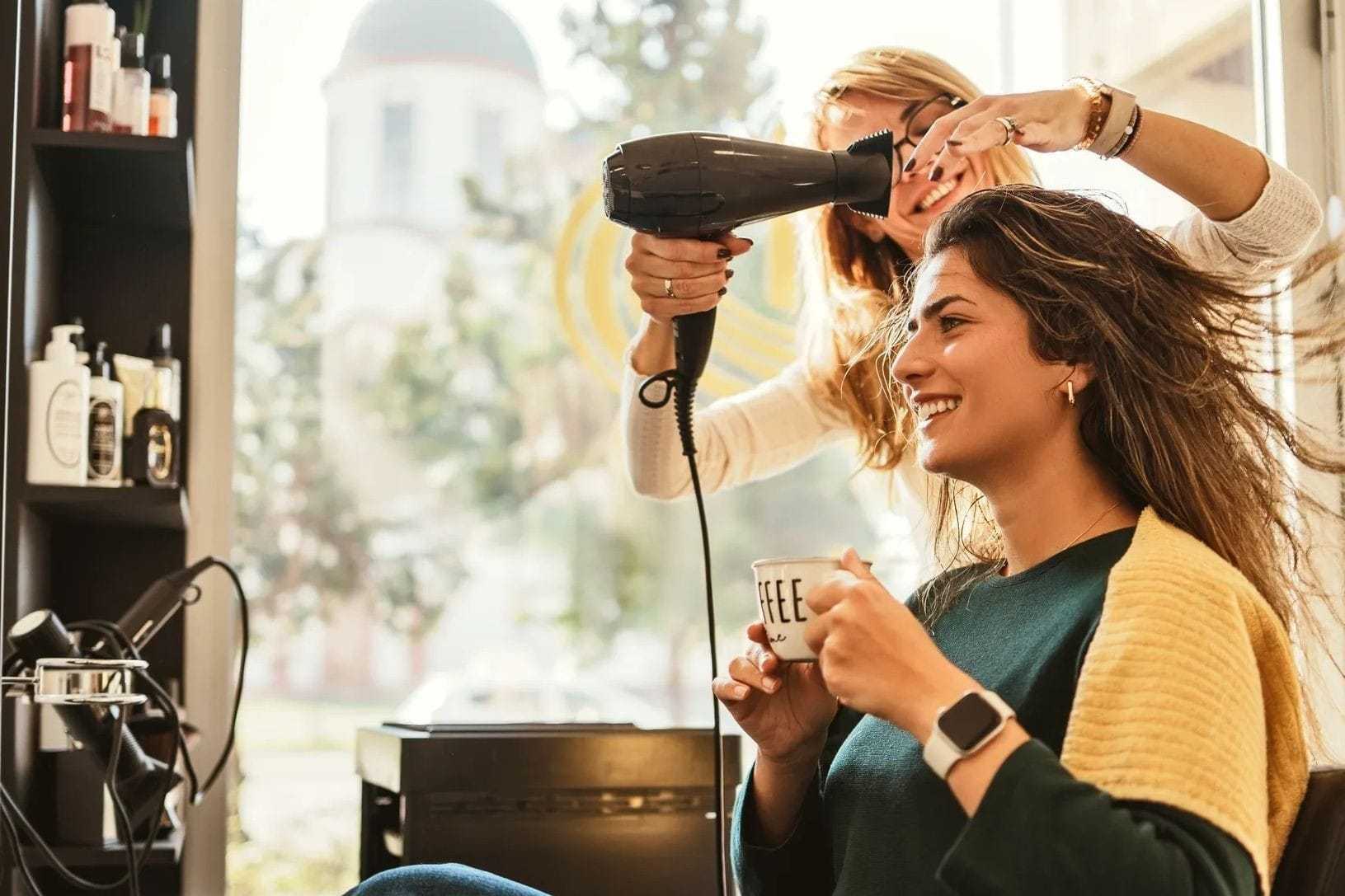Smiling woman gets hair styled while holding a coffee mug in a salon.