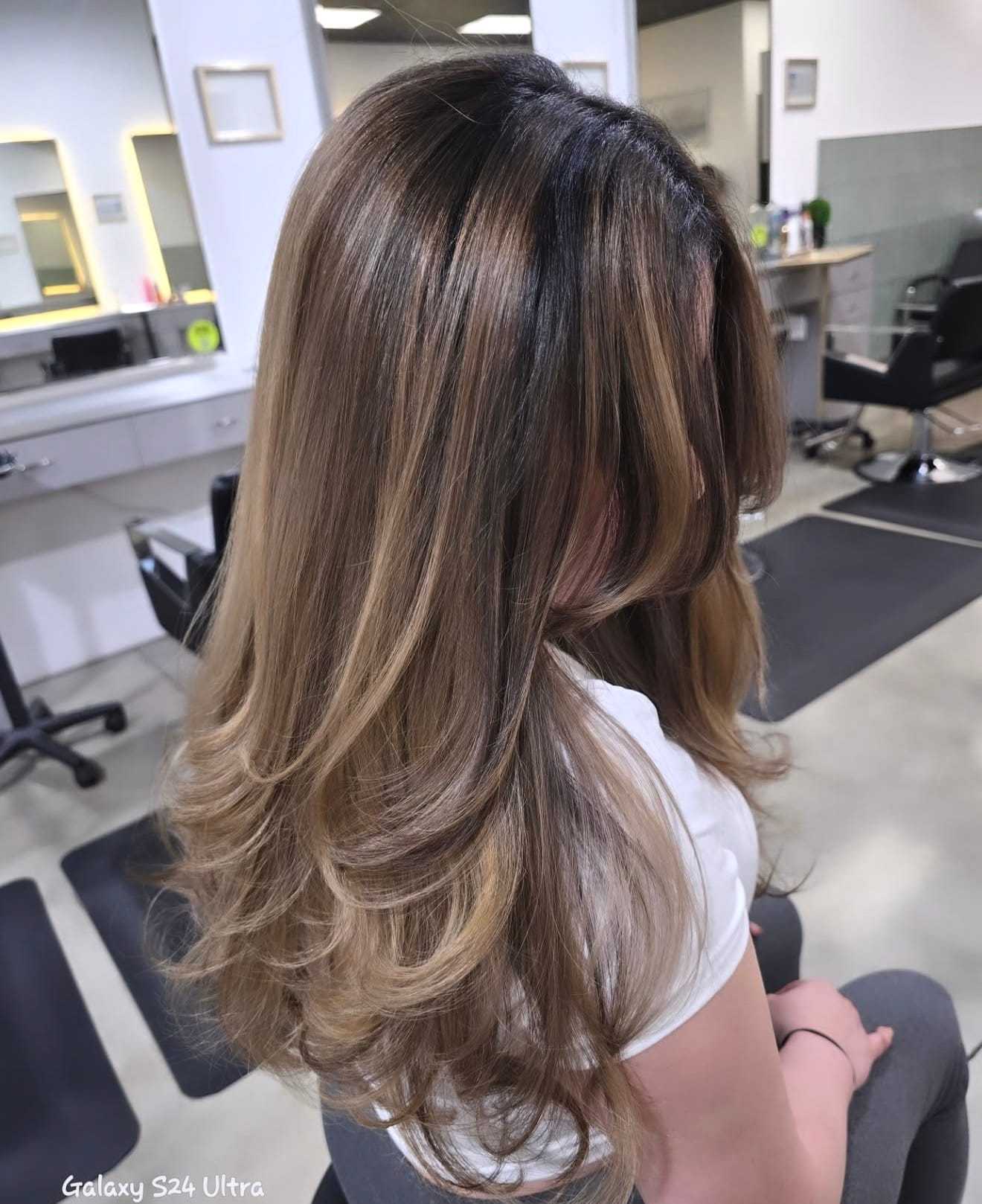 Hair salon interior with a person showing off long, layered brown hair with blonde highlights.