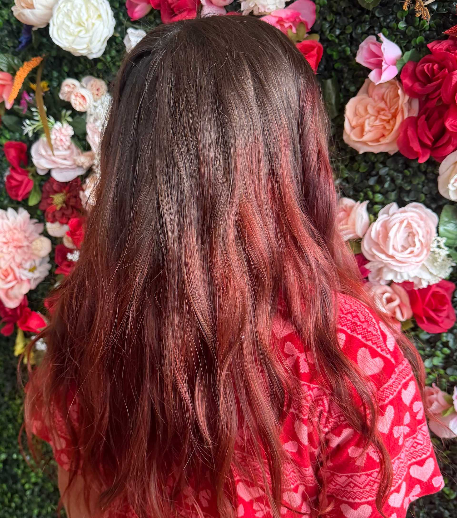 A person with long red hair stands in front of a vibrant floral backdrop.