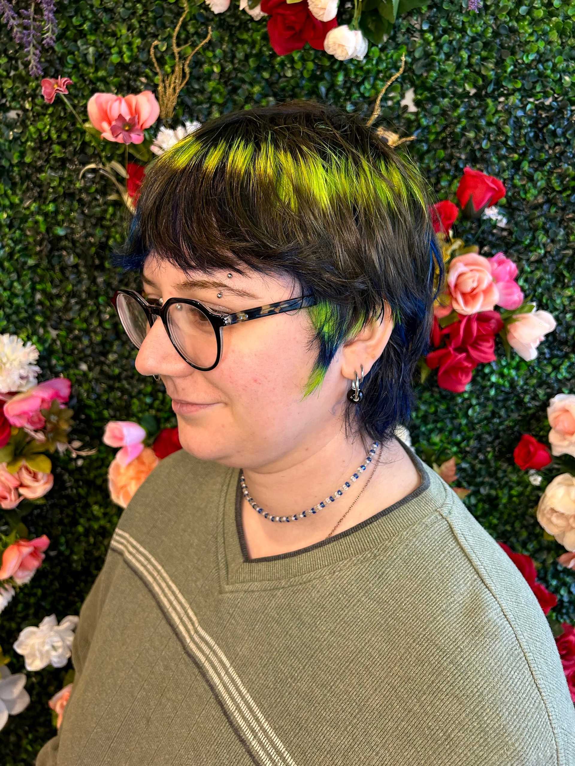 Person with glasses and colorful hair stands against a floral wall wearing a green sweater.